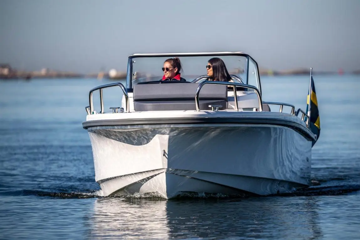 Two women enjoy a boat ride on calm waters, with a clear sky and a Swedish flag visible.